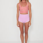 Woman wearing a red and white checkered punchy surf crop top and pink high-waisted betty bottoms 2.0 shorts on a white background