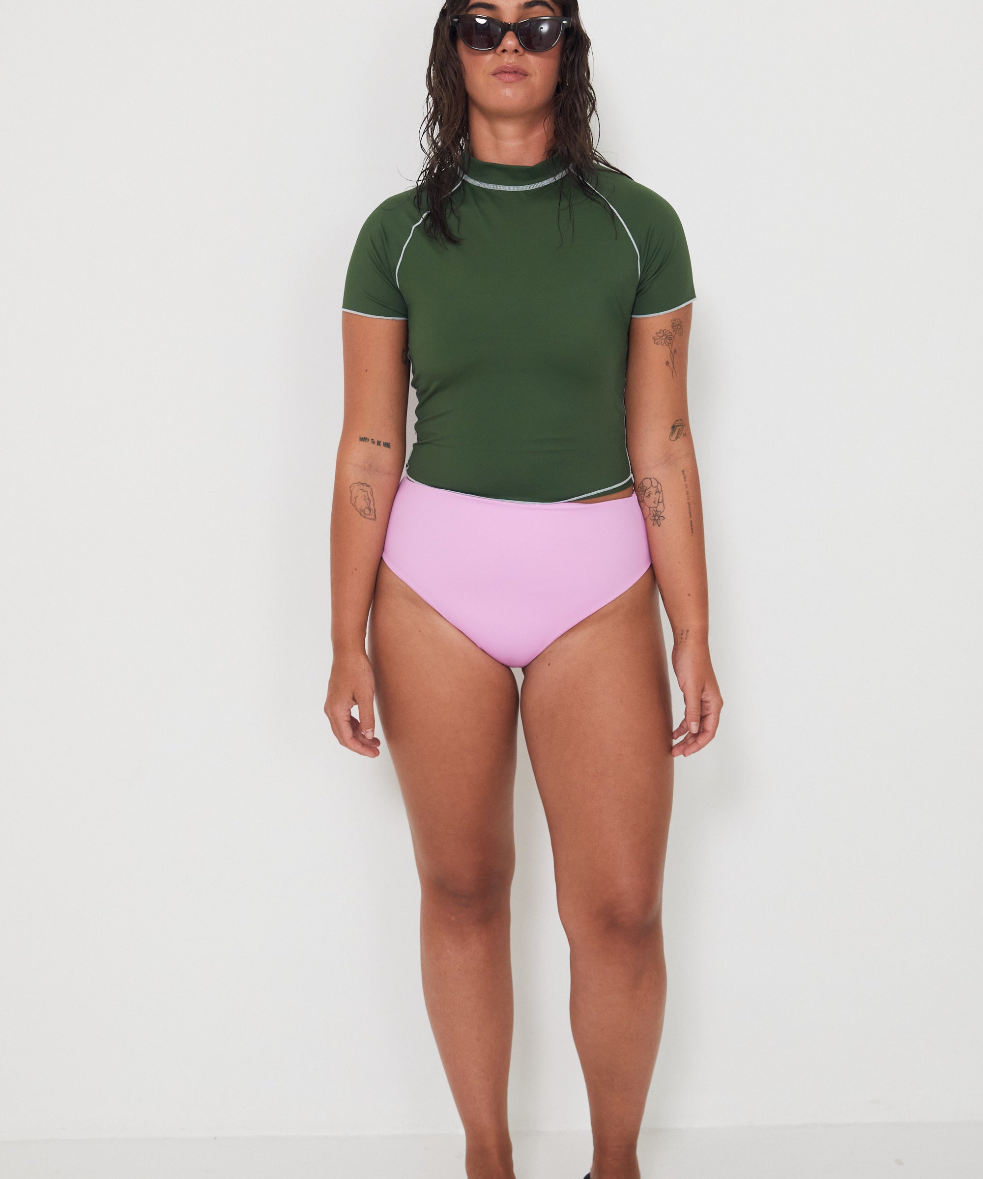 Woman wearing a green olive low rider rashguard and pink drifter swim bottoms with white trim on a plain background