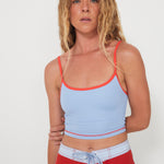 Woman wearing a light blue tankini surf top bra and red boarshorts on a white background