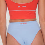 Person wearing a red 'Salt Gypsy' punchy surf bikini top and light blue drifter bottoms on a plain background