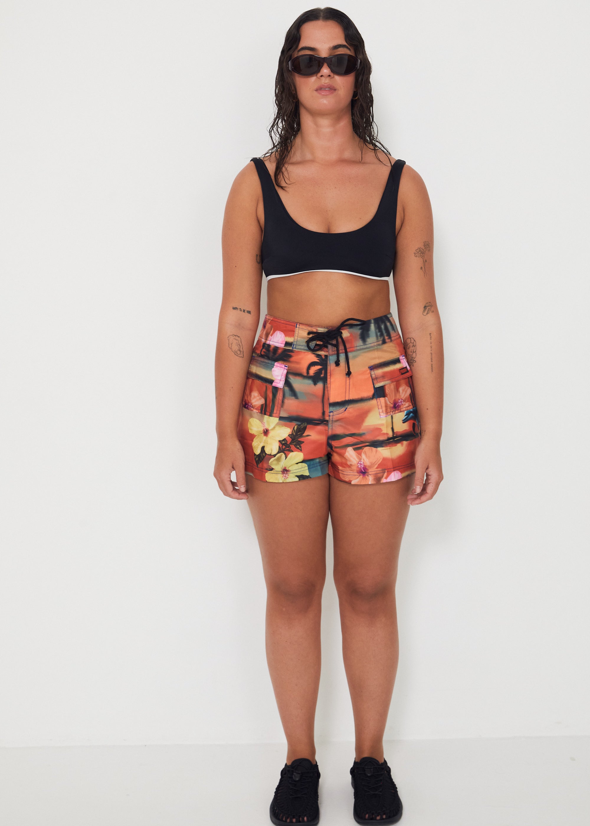 Woman wearing a black bralette swimwear crop top and vacay floral holiday swimwear boardshorts on a white background collaboration of salt gypsy and friday 