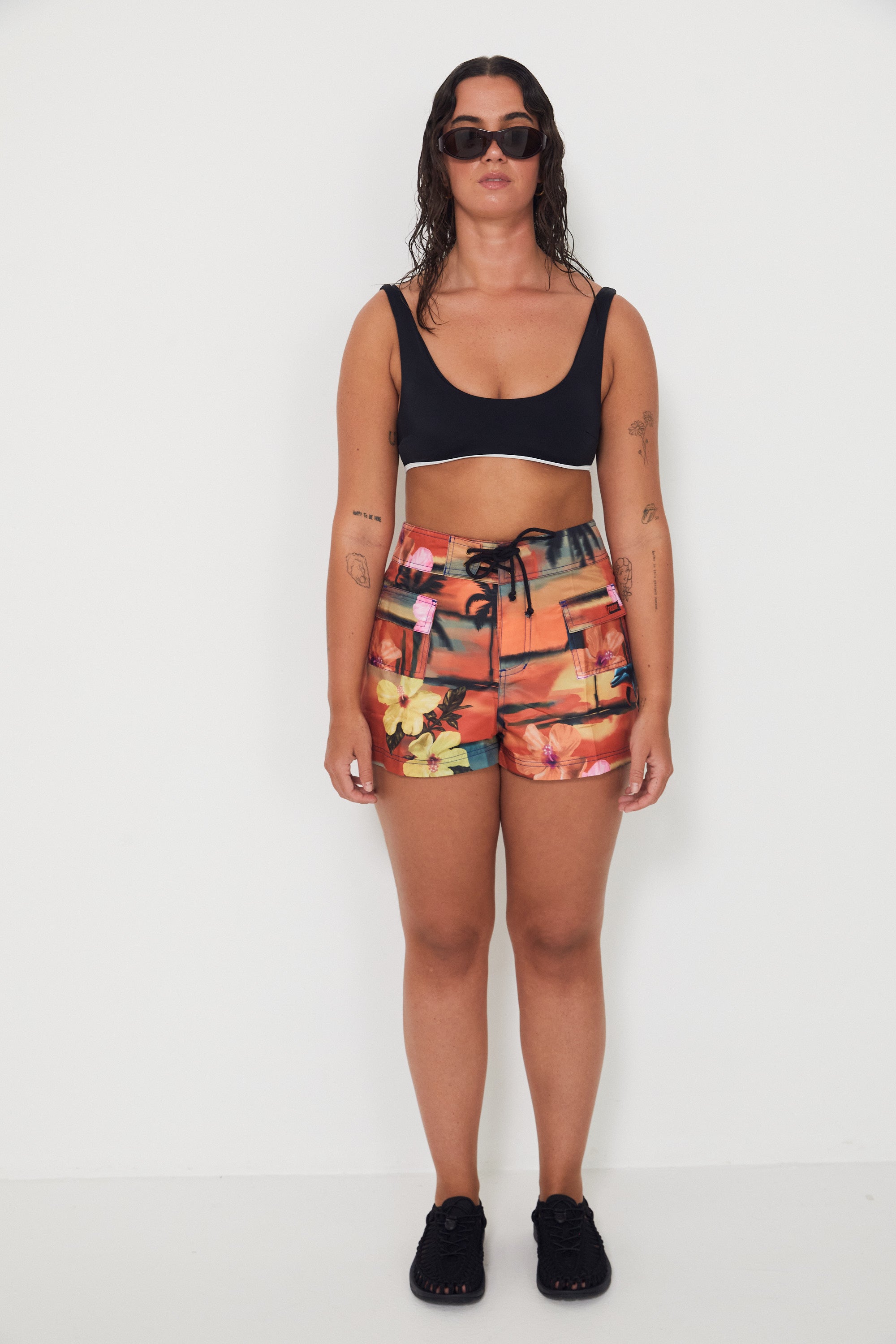 Woman wearing a black bralette swimwear crop top and vacay floral holiday swimwear boardshorts on a white background collaboration of salt gypsy and friday 