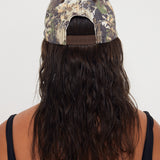 Person wearing a camouflage dad cap with long beachy hair flowing out of it on a white background