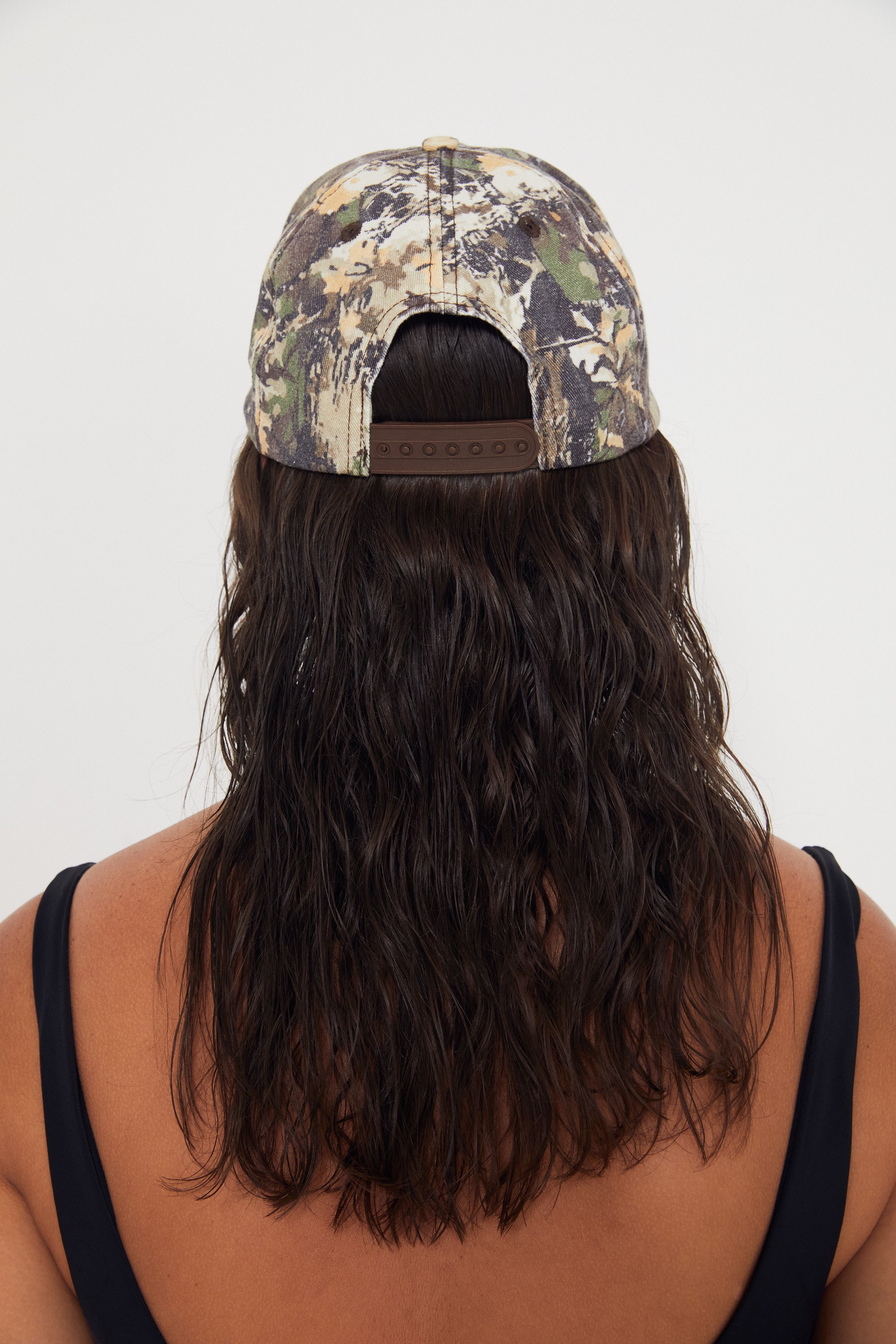 Person wearing a camouflage dad cap with long beachy hair flowing out of it on a white background